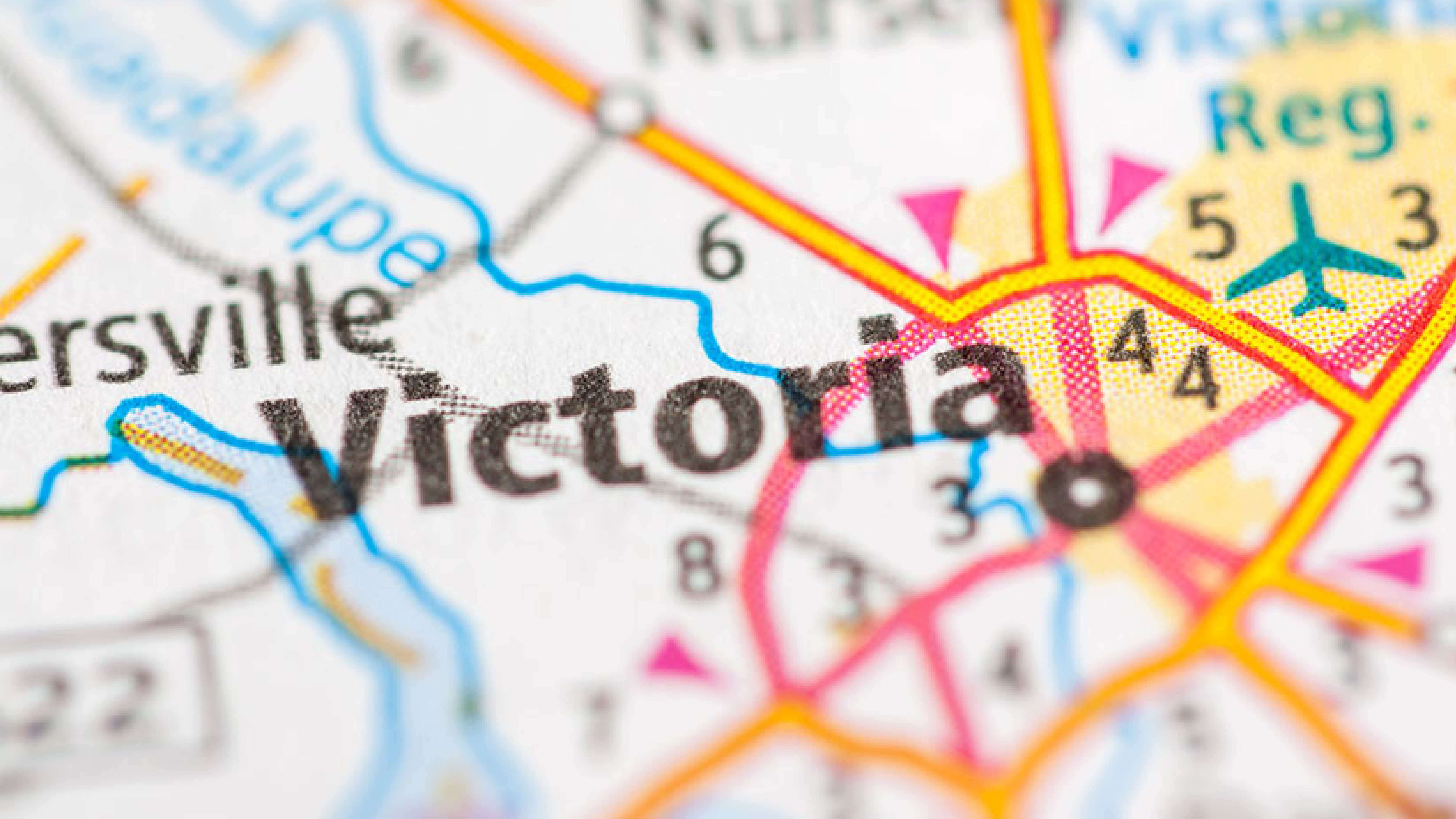 A close-up of a printed map highlighting Victoria, Texas, with surrounding roads, highways, and nearby cities partially visible. Bold black text marks "Victoria" at the center, with color-coded lines and icons indicating infrastructure like an airport and regional routes.